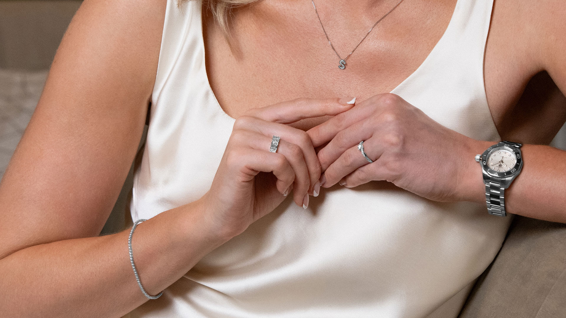 A female model wears an initial 'S' pendant and diamond tennis bracelet in 9ct white gold, with two platinum diamond rings and a TAG Heuer Aquaracer watch.